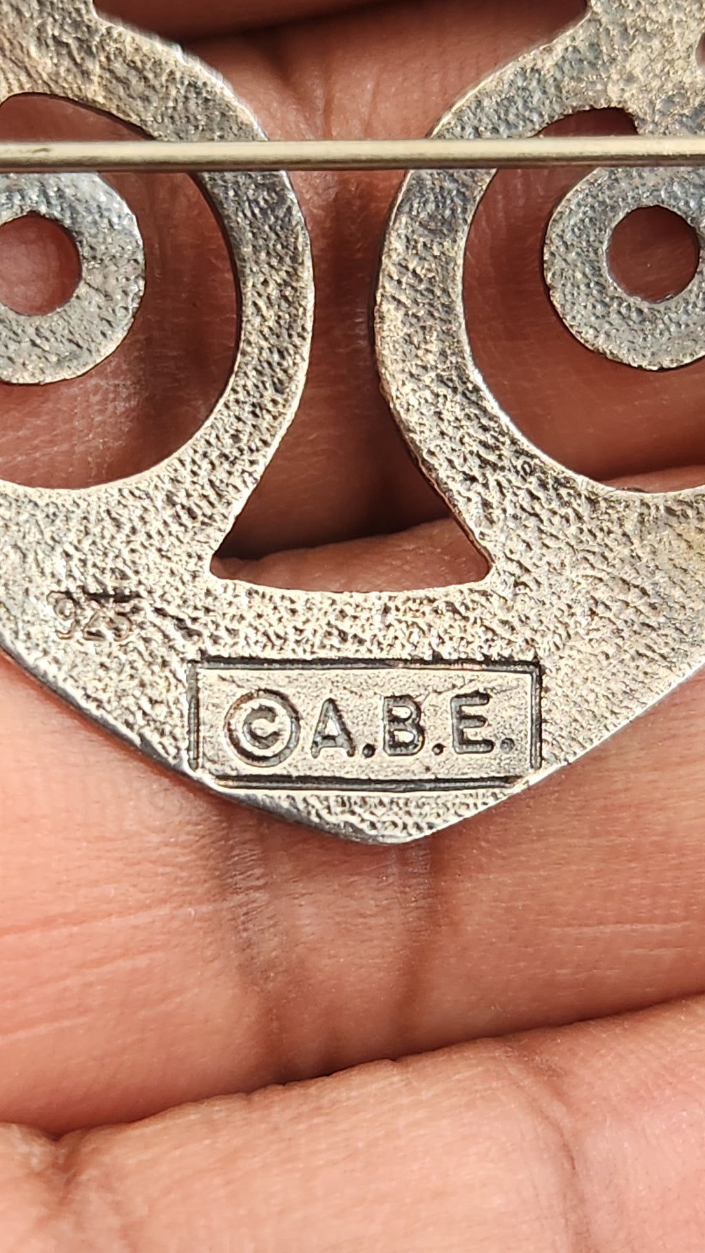 A close-up of a textured silver-toned metal accessory held in a person's hand, featuring cutout circular shapes and an embossed 'ABE