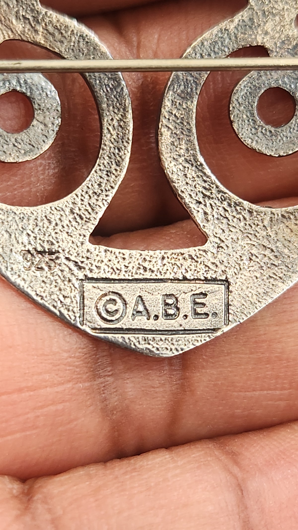 A close-up of a textured silver-toned metal accessory held in a person's hand, featuring cutout circular shapes and an embossed 'ABE