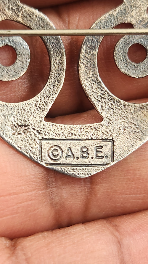A close-up of a textured silver-toned metal accessory held in a person's hand, featuring cutout circular shapes and an embossed 'ABE